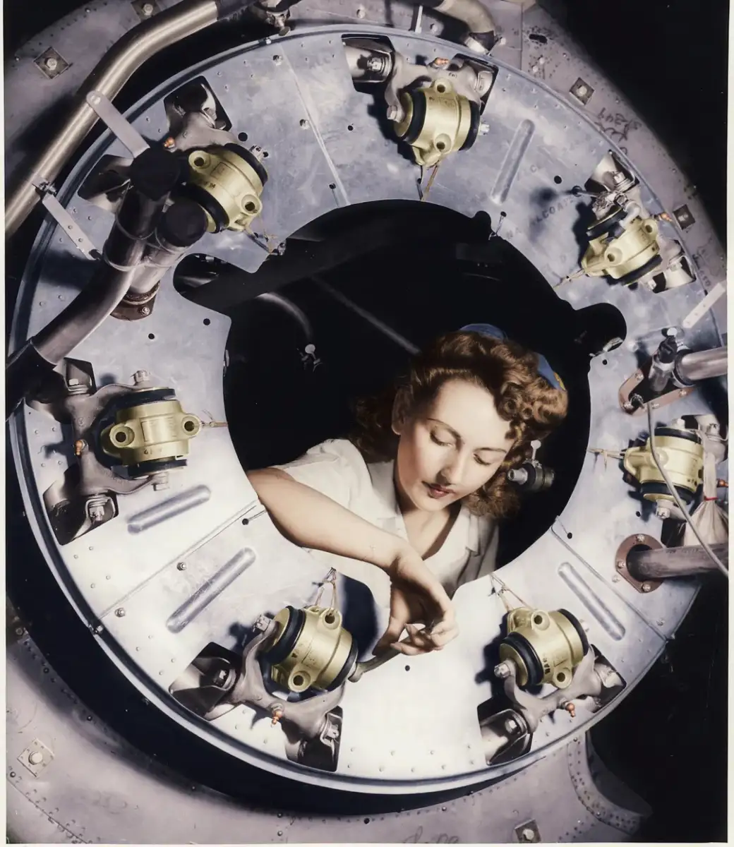A woman with curled hair and a white blouse works inside a large circular metal aircraft engine component, surrounded by mechanical parts and pipes.