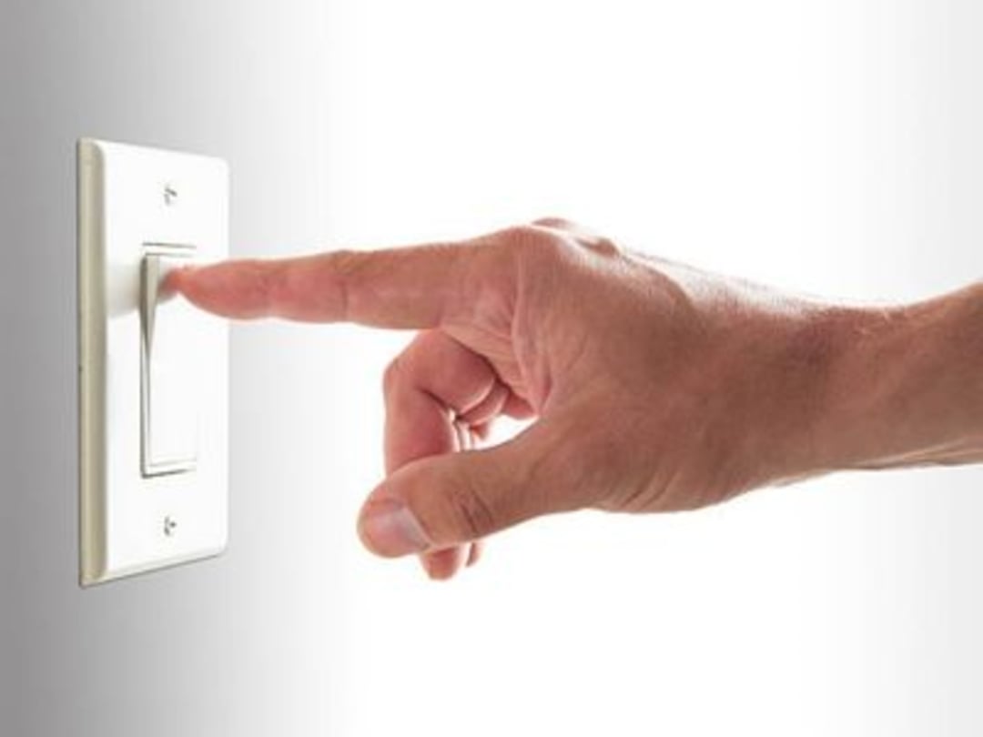 A hand reaching out with an extended index finger, about to press a light switch on a white wall.