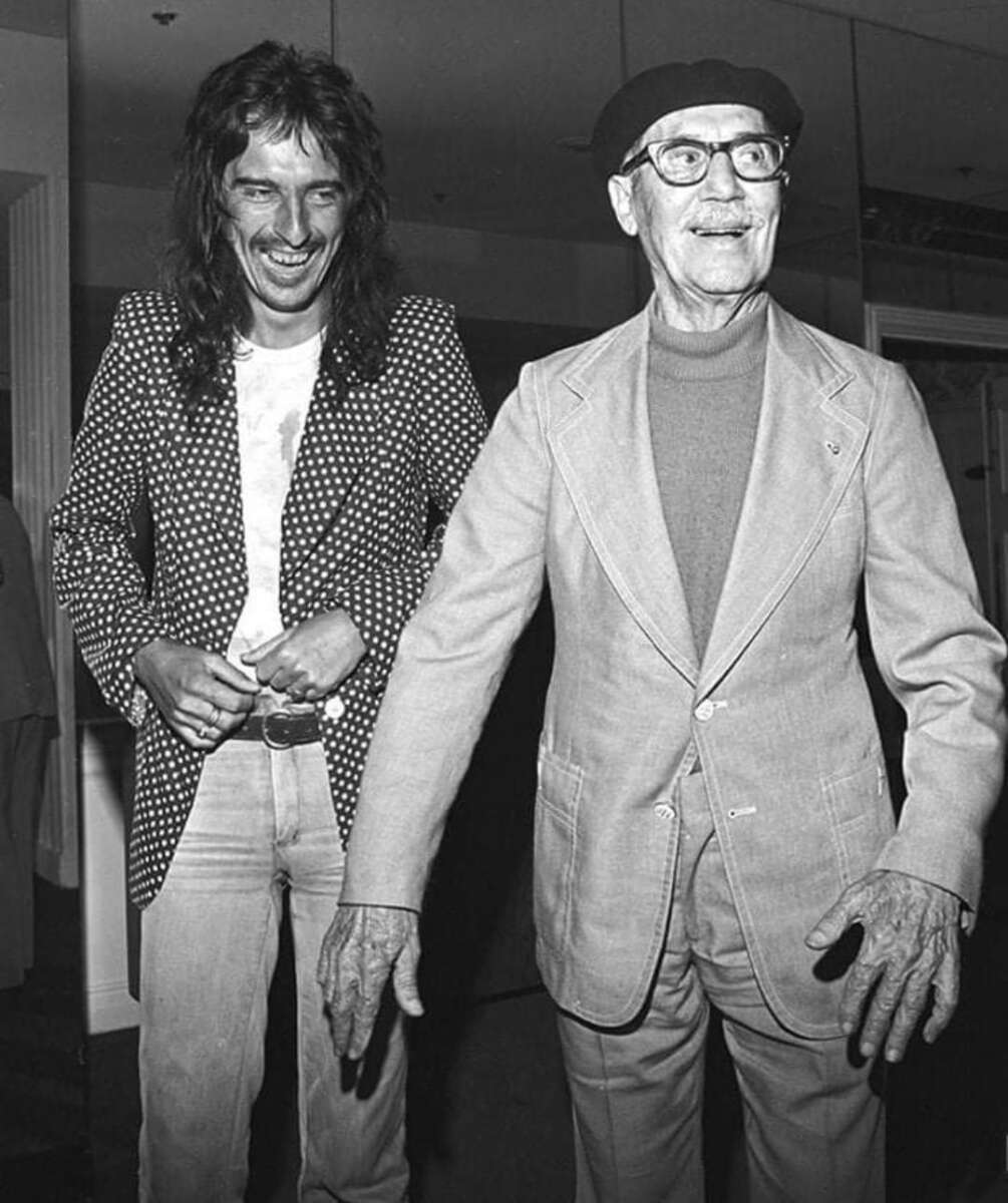 Two men stand together, smiling. The man on the left has long hair, wears a polka-dot jacket and jeans; the man on the right wears glasses, a beret, and a light-colored suit. Both appear happy and relaxed.