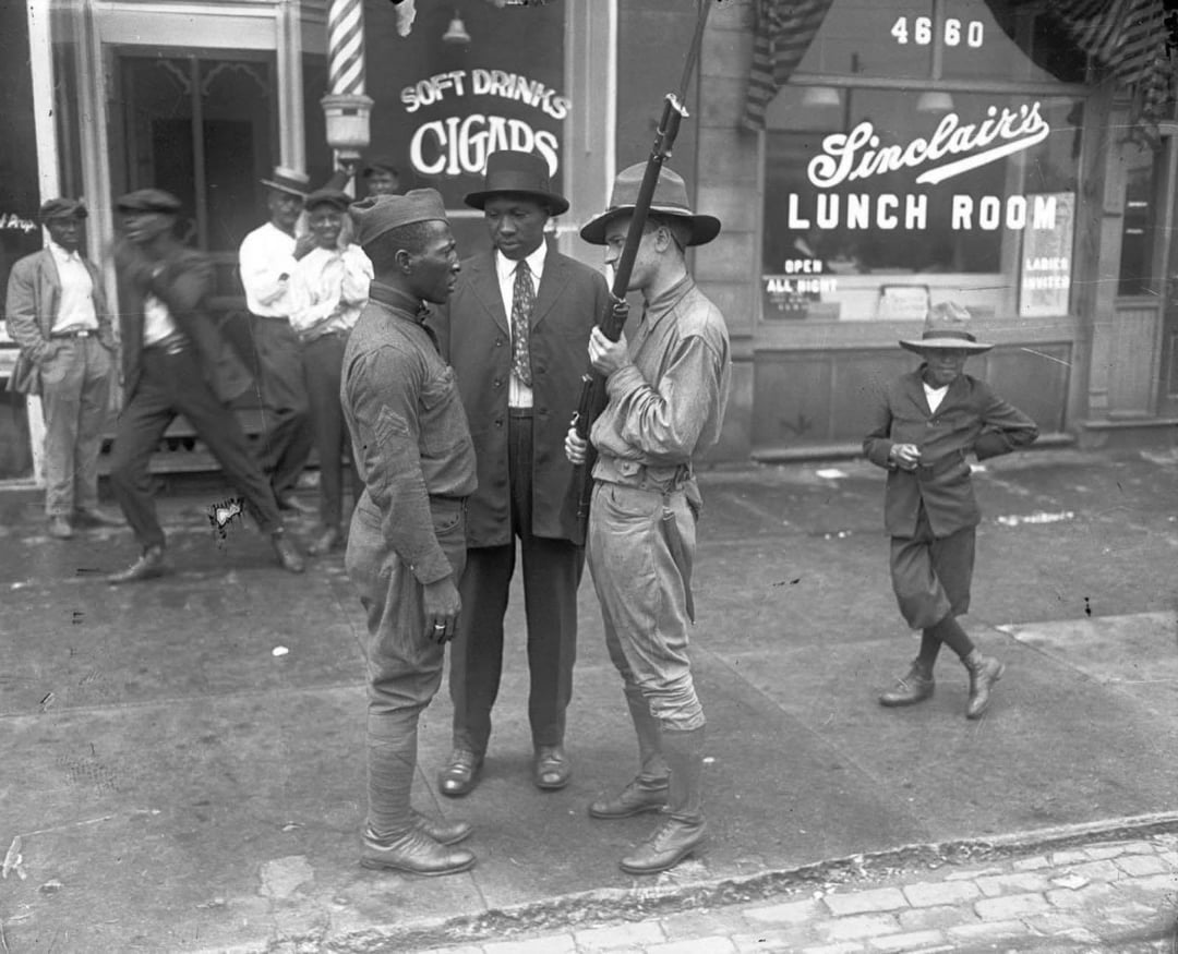 A Black soldier stands face-to-face with a white soldier holding a rifle on a city sidewalk, while men in suits and a boy stand nearby in front of shops labeled “Sinclair’s Lunch Room” and “Soft Drinks Cigars.”