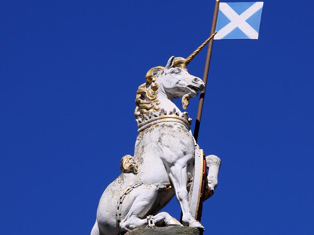 A white unicorn statue with a golden mane and horn holds a shield and a pole bearing the Scottish flag, set against a clear blue sky.