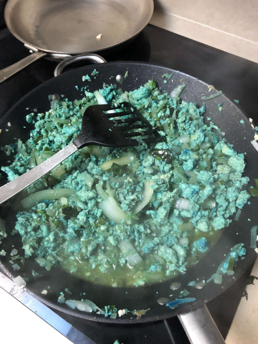 Ground meat and chopped onions are being cooked in a pan on a stovetop. The meat is an unusual bright blue-green color, and a black spatula rests in the pan. Steam is rising from the mixture.