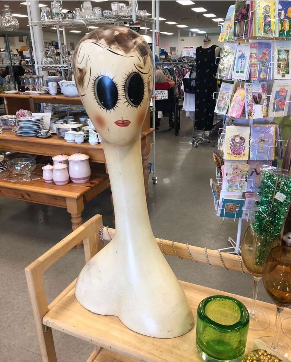 A mannequin head with exaggerated large painted eyes and lips, a long neck, and short painted hair stands on a cart in a thrift store filled with dishes, glassware, and greeting cards.
