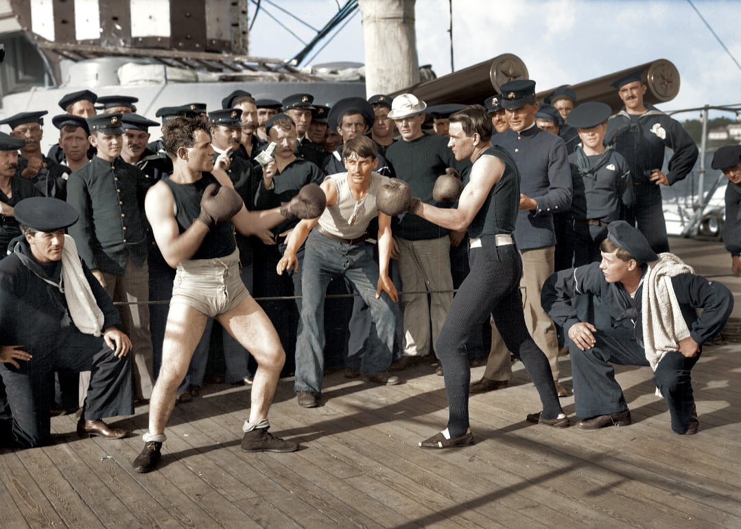 Two men wearing boxing gloves and old-fashioned athletic gear prepare to box on a ship’s deck, surrounded by sailors in navy uniforms watching and cheering them on.