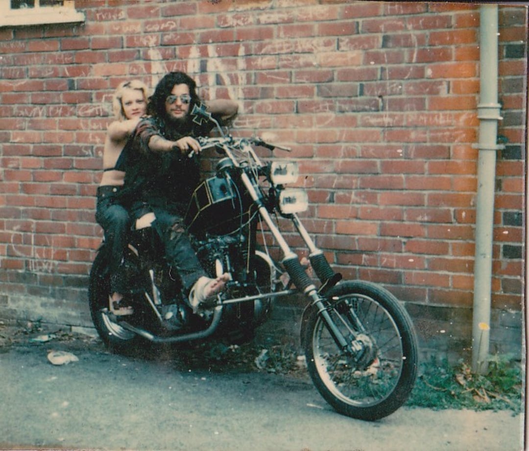 Two people sit on a custom motorcycle in front of a red brick wall. One person has long dark hair and sunglasses; the other has short blond hair and an arm around the first person's shoulders. Both appear relaxed and casual.