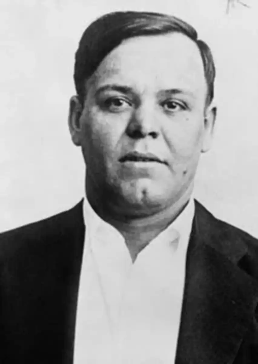 Black and white portrait of a man with short dark hair, wearing a dark jacket and a white shirt, looking directly at the camera with a neutral expression.
