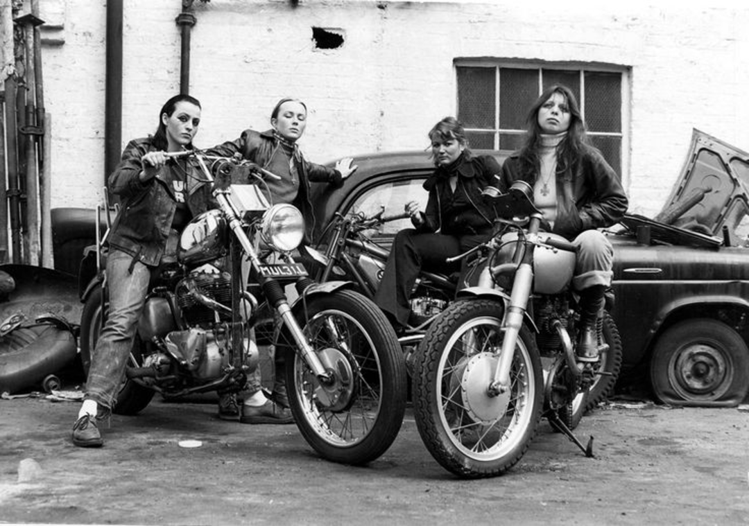 Four women in leather jackets pose confidently with motorcycles in front of an old car and a brick wall in a gritty, urban setting. Two women are seated on motorcycles, while the other two stand beside them.
