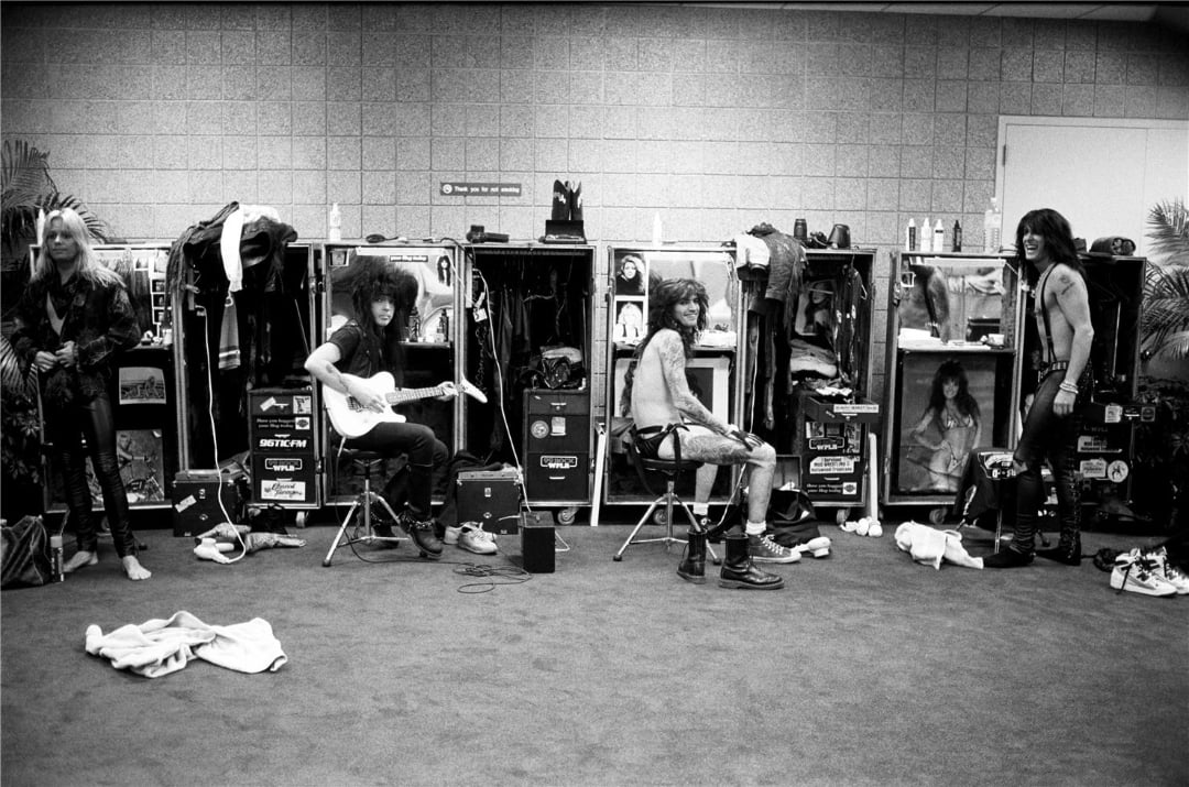Five people in a backstage room sit or stand near open lockers filled with clothes and equipment; one plays an electric guitar while others relax or prepare, with scattered items on the floor.