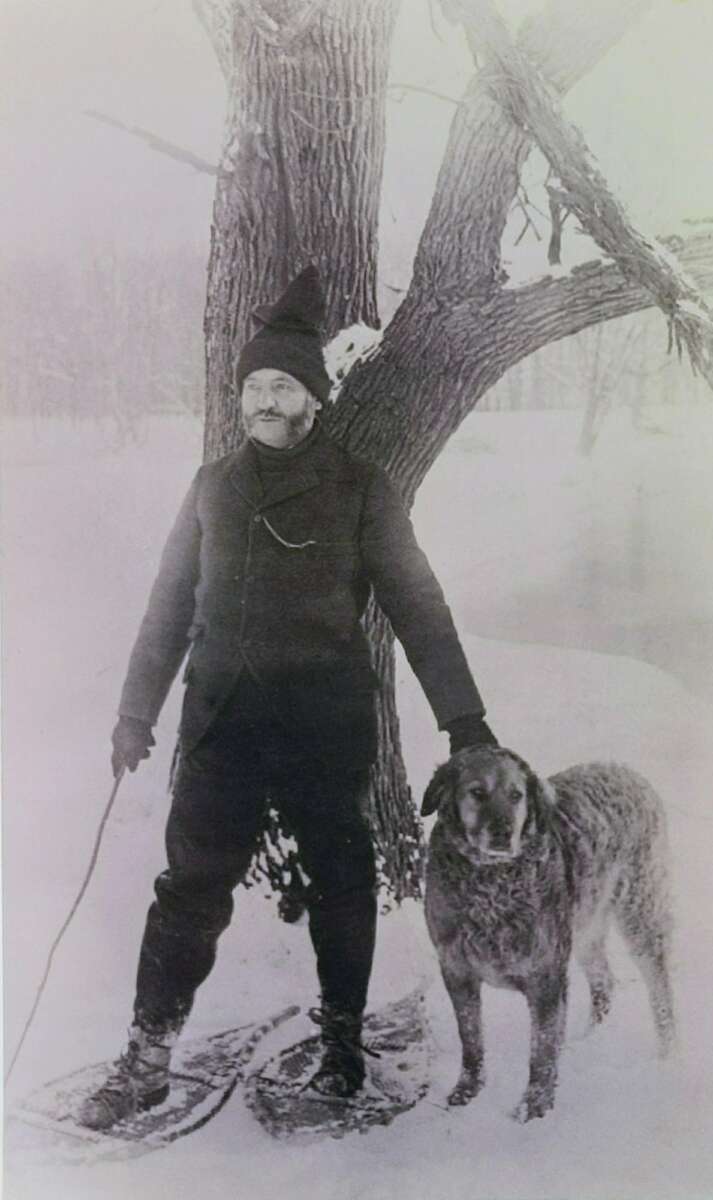 A man wearing winter clothing and snowshoes stands beside a large dog in front of a tree on a snowy landscape. The man holds a walking stick and rests his hand on the dog.