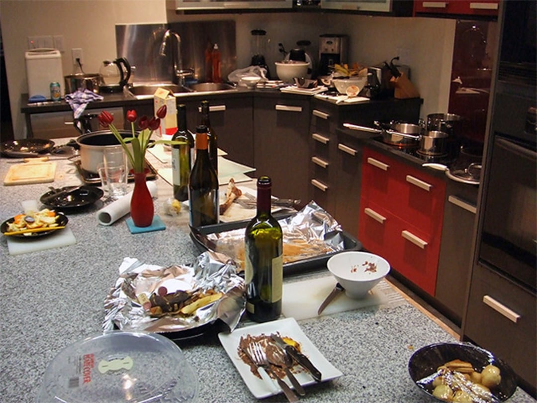 A modern kitchen with messy countertops covered in dirty dishes, food scraps, empty wine bottles, and baking trays. A vase with red tulips sits among the clutter, and pots and pans pile up near the sink.