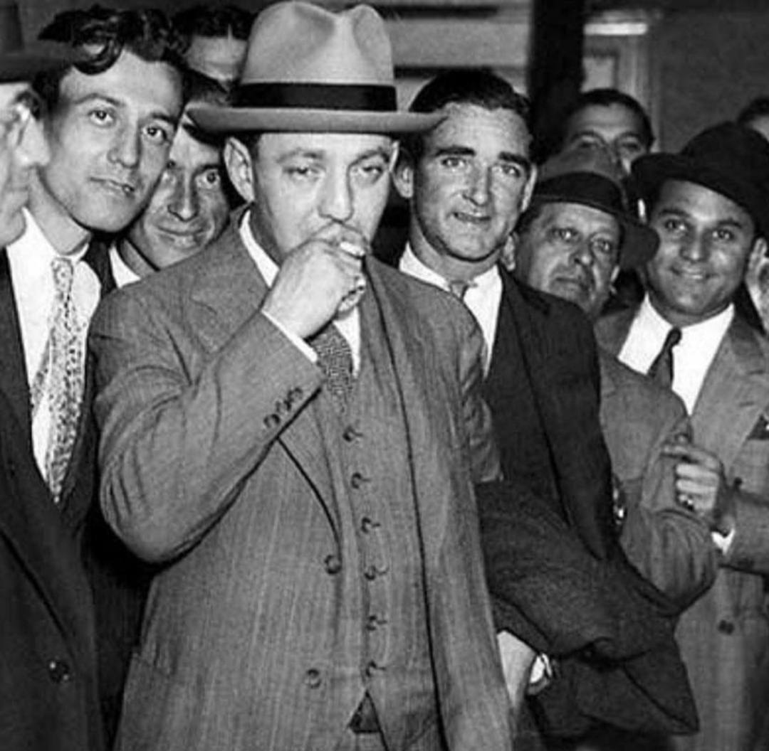 A group of men in suits and hats stand closely together indoors. The man in front wears a three-piece suit and fedora, with his hand near his mouth, while others behind him look on and smile. The photo is in black and white.
