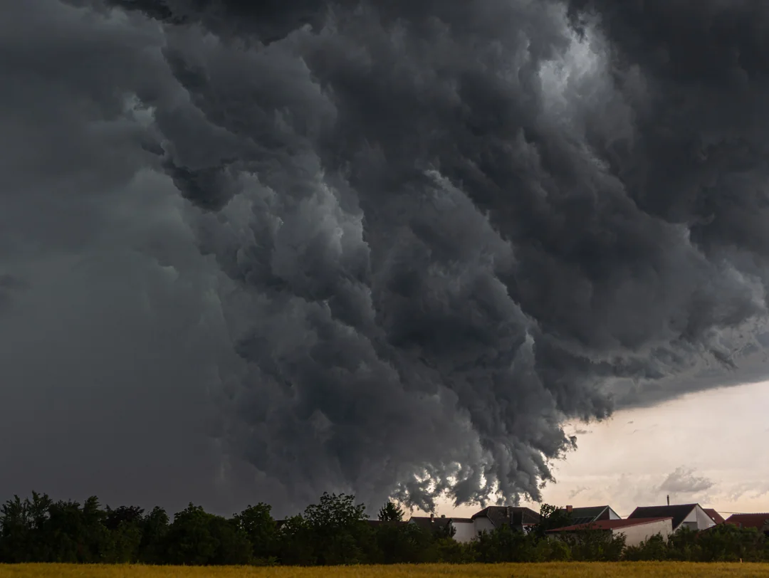 Dark, dramatic storm clouds loom over a small village with houses and trees, creating a striking contrast between the turbulent sky and the calm landscape below.