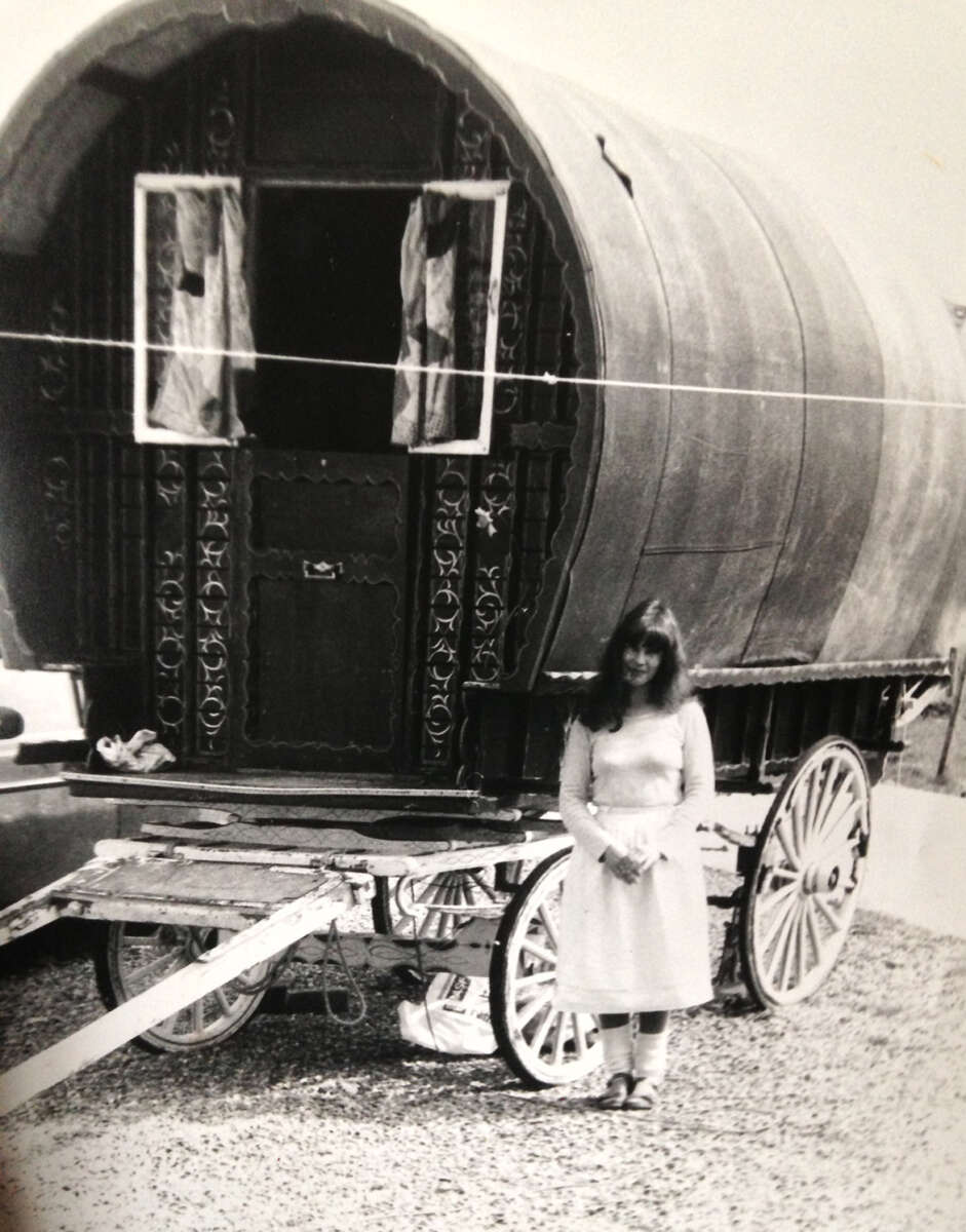 A young woman in a light-colored dress stands in front of a traditional, ornate wooden wagon with a curved roof and decorative details. The photo is black and white, evoking a vintage feel.
