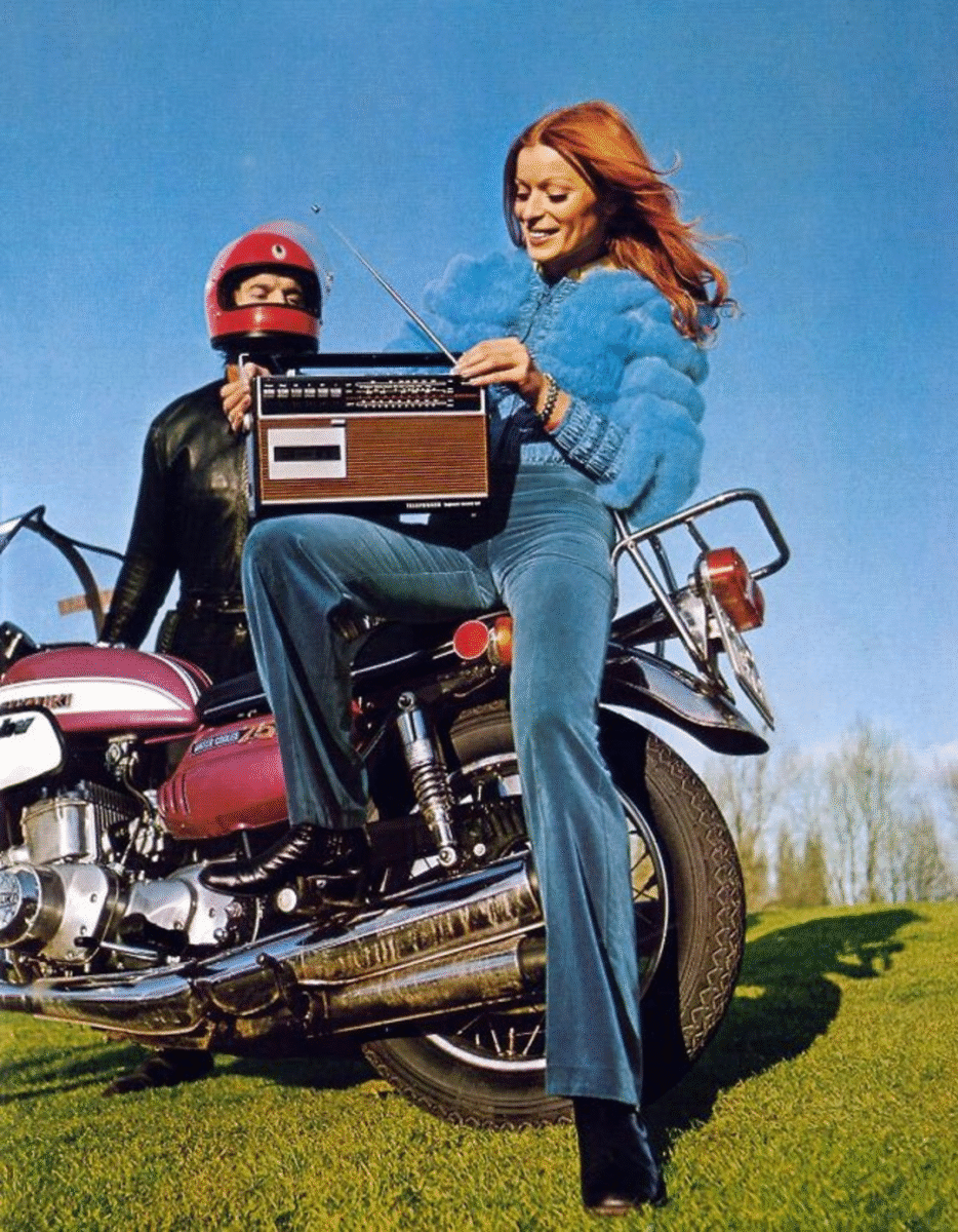 A smiling woman in a blue outfit sits on a motorcycle holding a vintage radio, while a person in a red helmet stands beside her. The sky is clear and the grass is green, suggesting a sunny day.