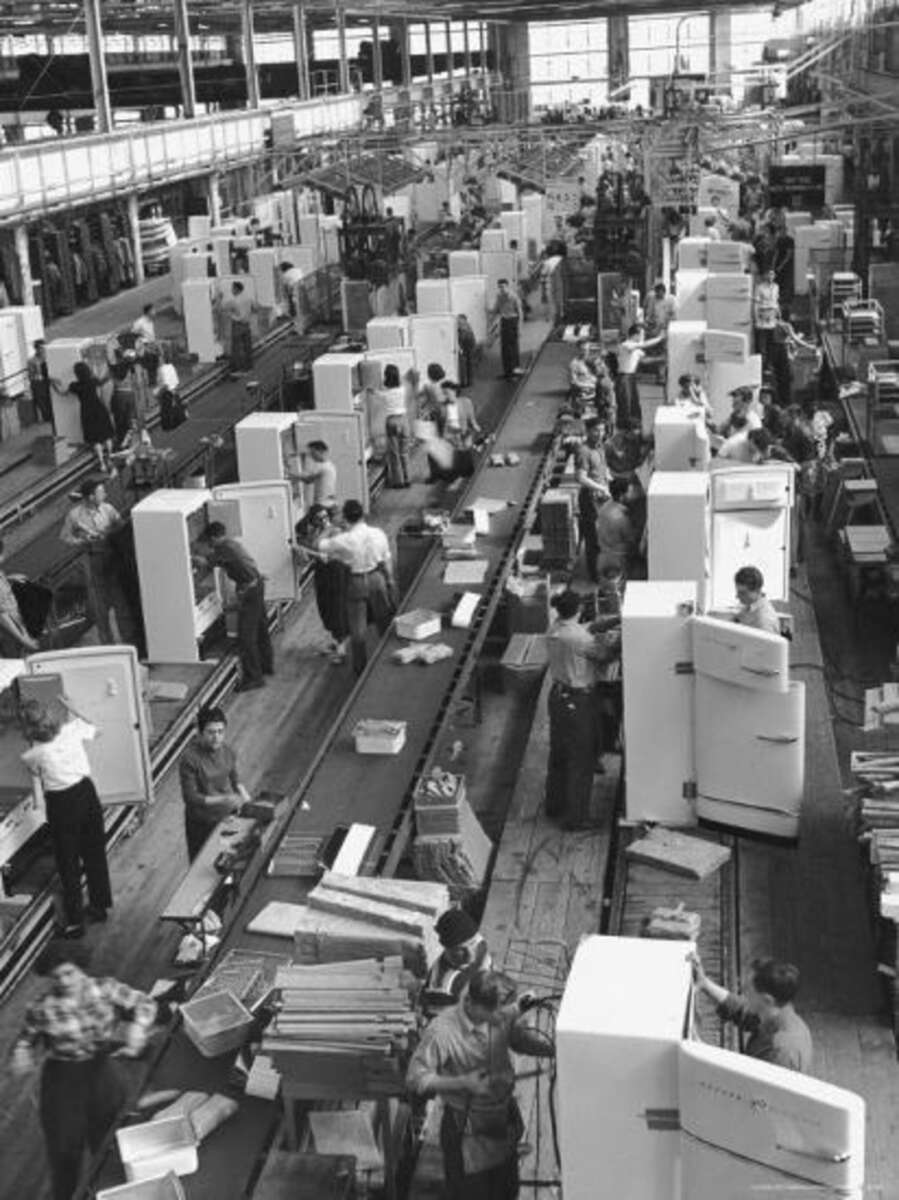 Black and white photo of a busy factory assembly line where workers are assembling refrigerators, with open refrigerator doors visible and people positioned on both sides of the conveyor belt.