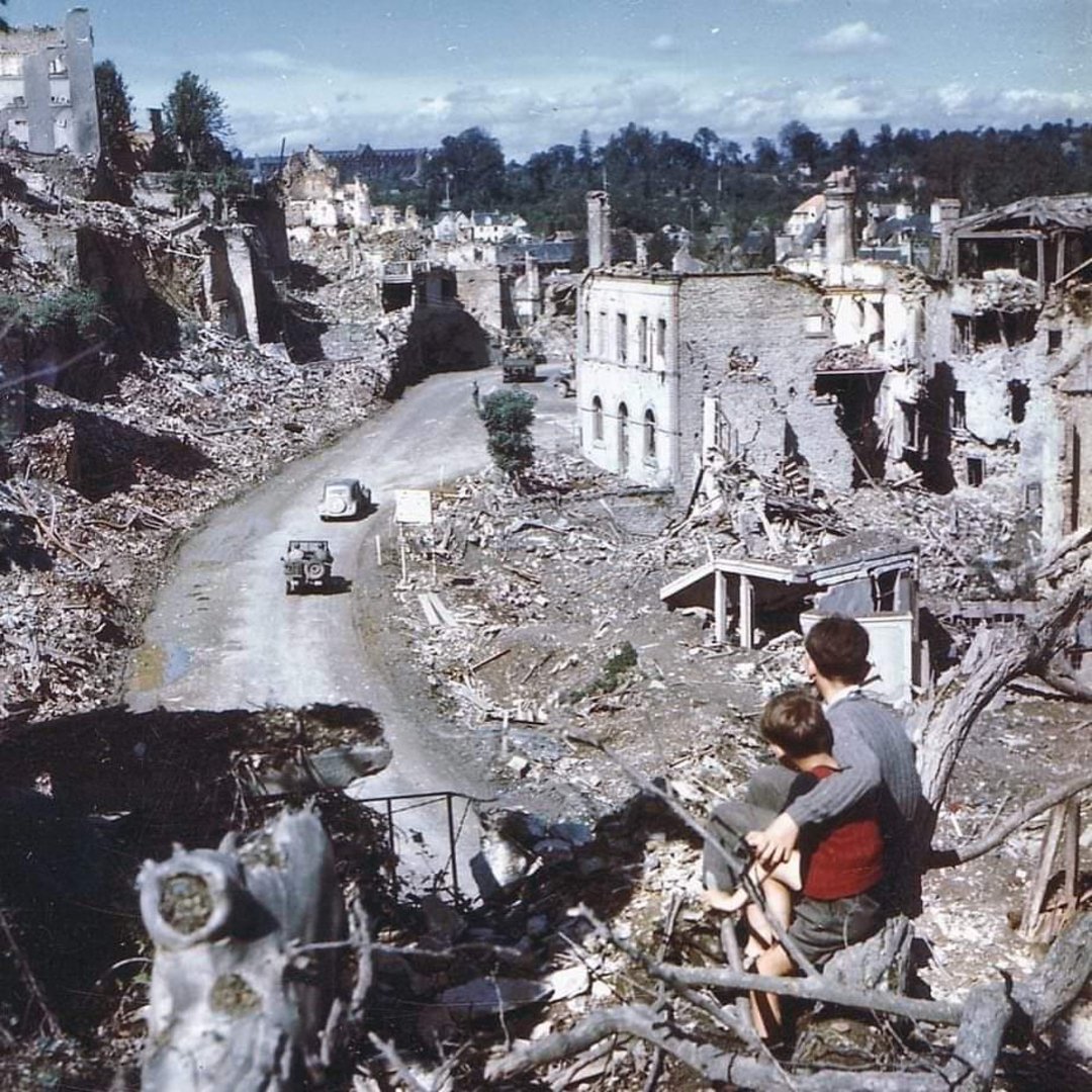 A man and child look over a devastated, rubble-filled street with destroyed buildings on both sides as military vehicles drive down the road in a war-torn landscape.