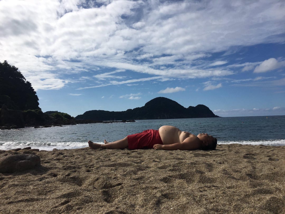 A person in red shorts lies on their back on a sandy beach near the shoreline, with the ocean and a tree-covered hill in the background under a partly cloudy sky.