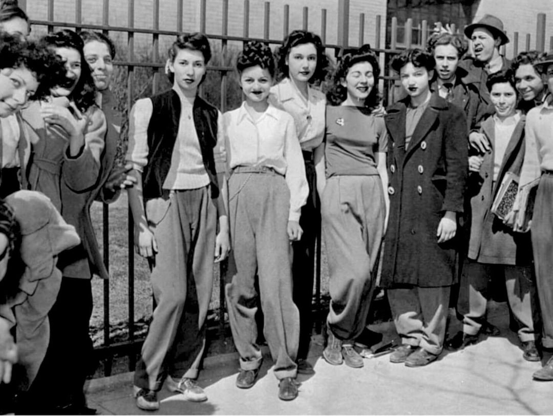 A group of young women and a few men stand and smile together in front of a metal fence, wearing 1940s-style clothing, including high-waisted pants, jackets, and coats, in a black-and-white outdoor photo.