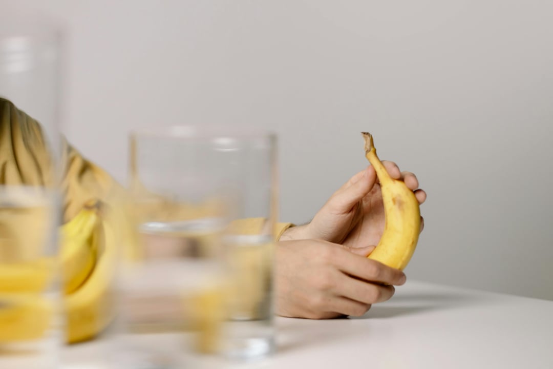 A person holding a ripe banana in one hand is seated at a white table with two clear glasses of water and another banana in the foreground. The background is plain and neutral.
