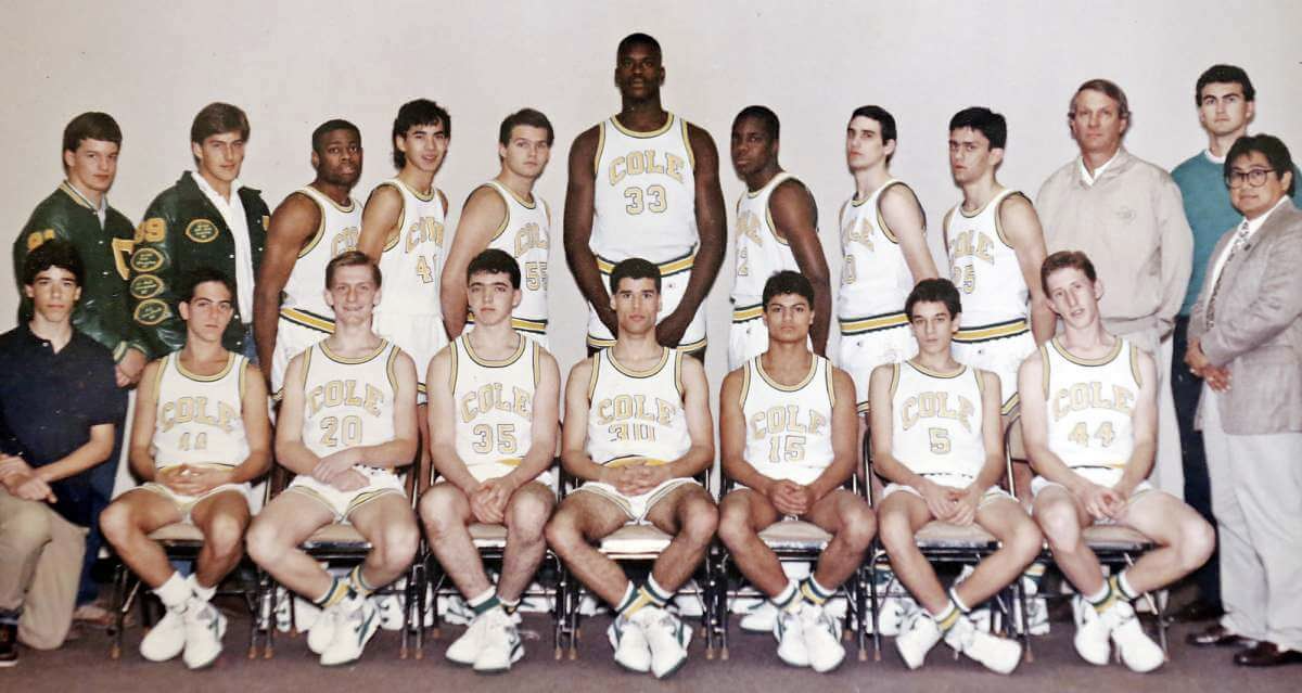 A high school basketball team poses for a group photo. Eight players in uniforms are seated in front, with more players, coaches, and staff standing behind them against a plain backdrop.