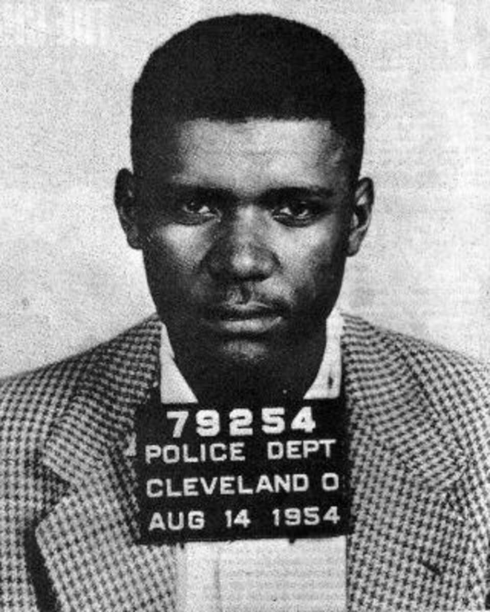 Black-and-white mugshot of a man in a checkered jacket holding a sign reading "79254 Police Dept Cleveland O Aug 14 1954." He is looking straight at the camera with a neutral expression.