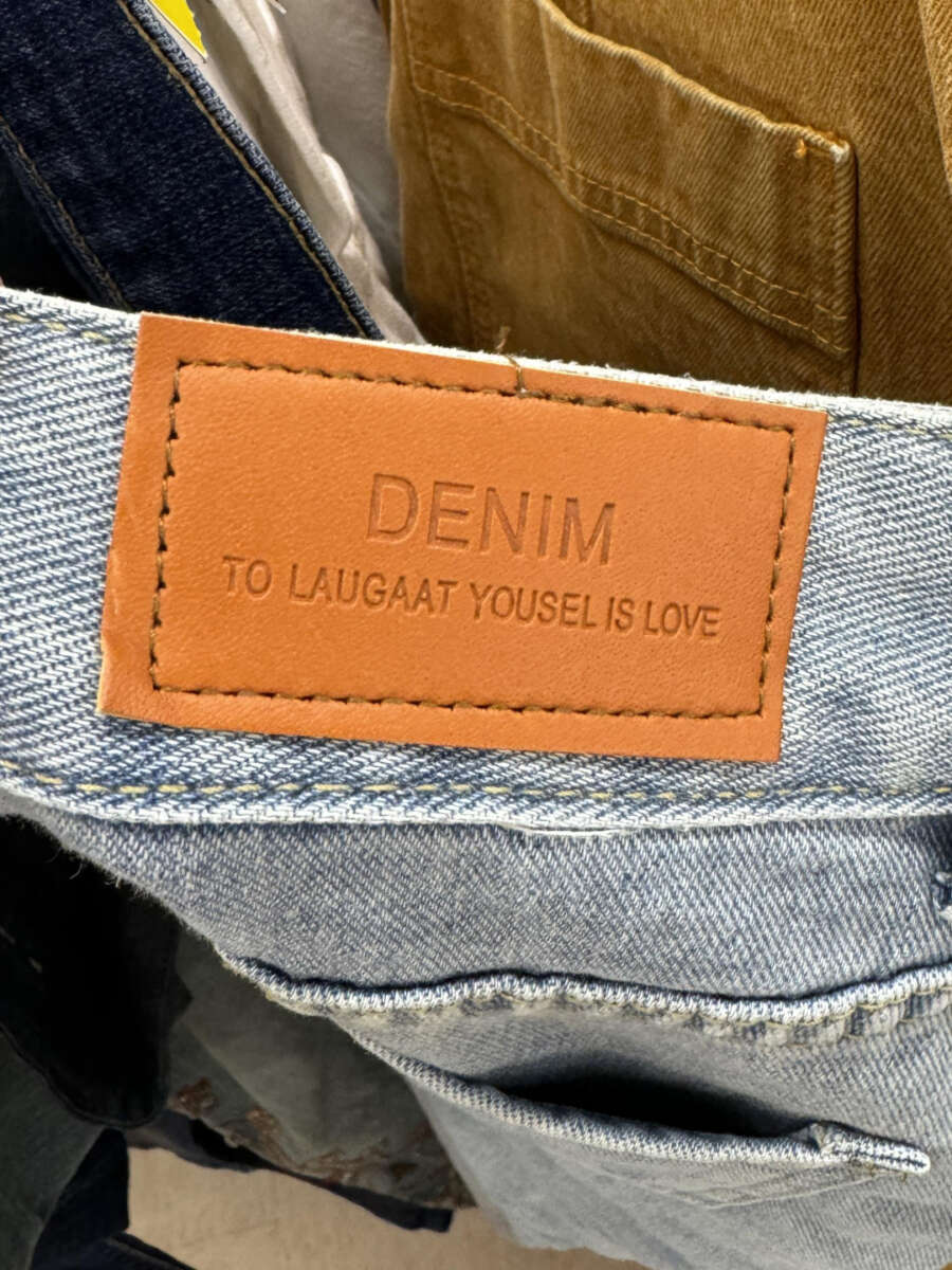 A close-up of a brown leather patch on light blue denim jeans, embossed with the words "DENIM TO LAUGAAT YOUSEL IS LOVE" in uppercase letters.