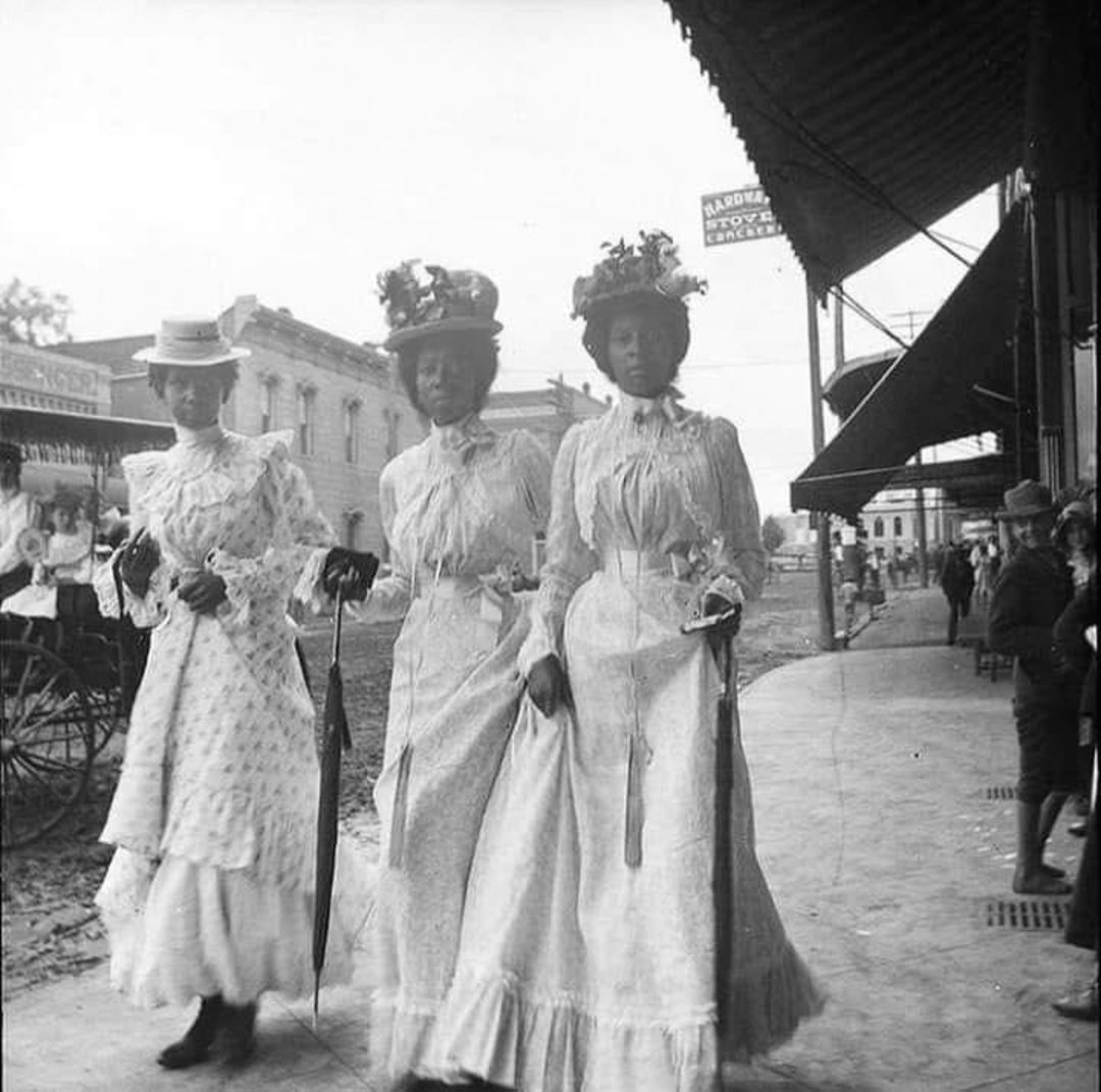 Three Black women in elegant long dresses and hats walk down a city sidewalk in an early 1900s street scene, with historic buildings, a horse-drawn carriage, and people in the background.