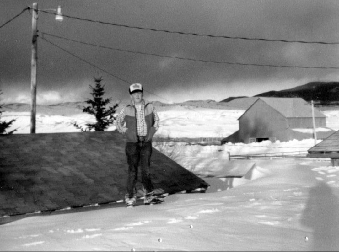 A person stands on deep snow next to a snow-covered rooftop. In the background are trees, utility poles with wires, distant houses, and a snowy landscape under a cloudy sky. The image is in black and white.