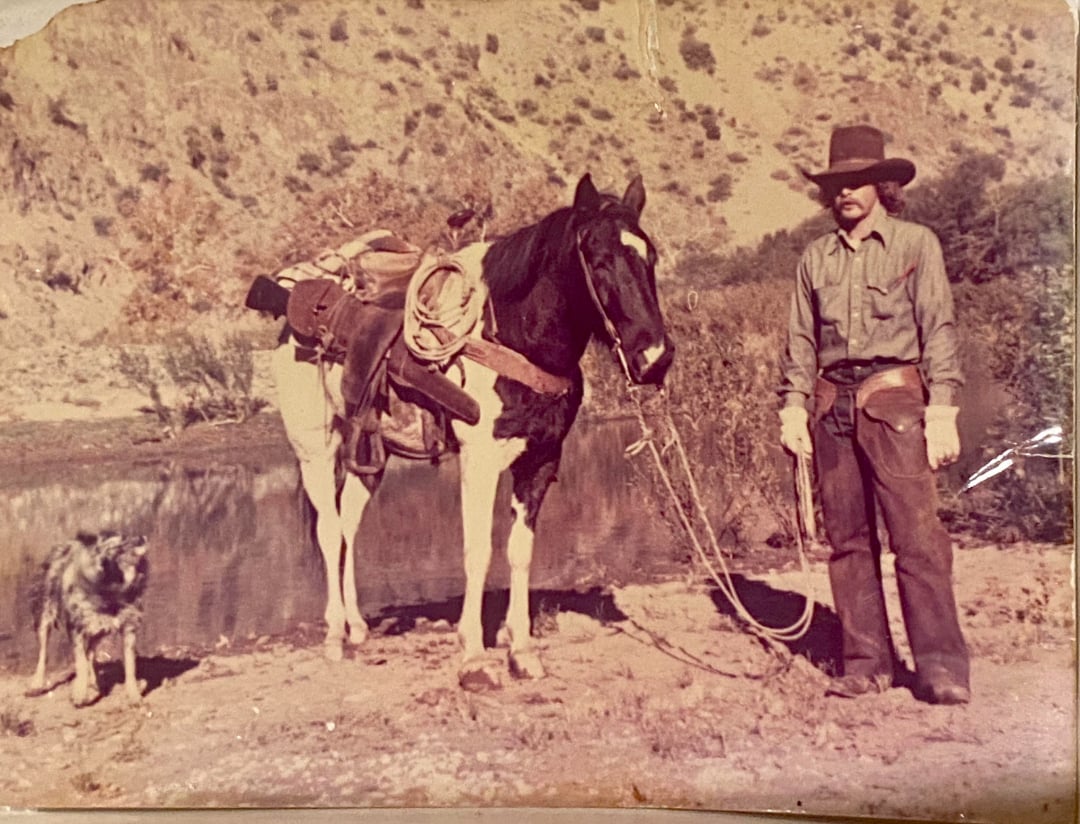 A man in cowboy attire stands next to a saddled horse by a riverbank, holding its reins. A dog stands nearby. The background features dry, brush-covered hills under bright sunlight. The photo appears vintage.