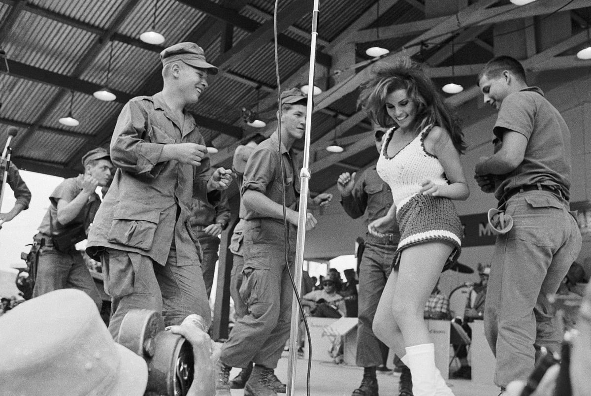 A woman in a crochet top and shorts dances with smiling soldiers in uniform under a metal-roofed structure, as others watch and clap in the background. A microphone is visible at the center.