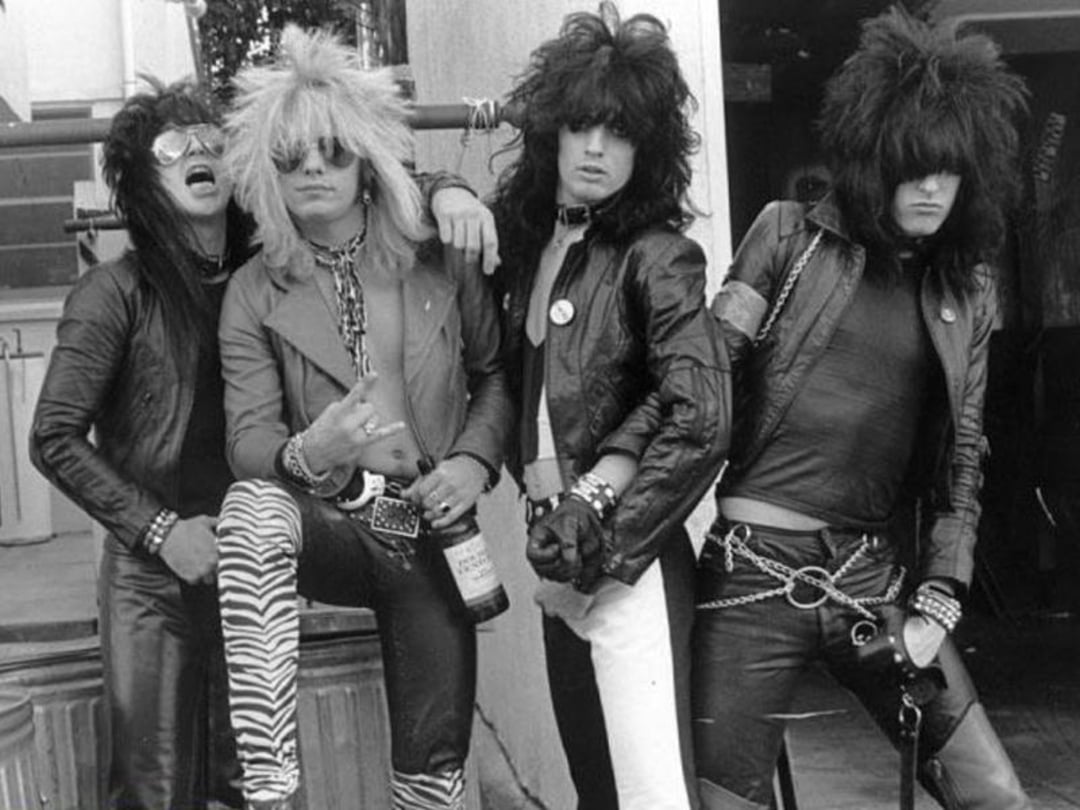 Four men with big, teased hair and 1980s glam rock outfits, including leather jackets, animal print pants, and sunglasses, pose together. One holds a bottle, standing confidently and expressing rebellious energy.