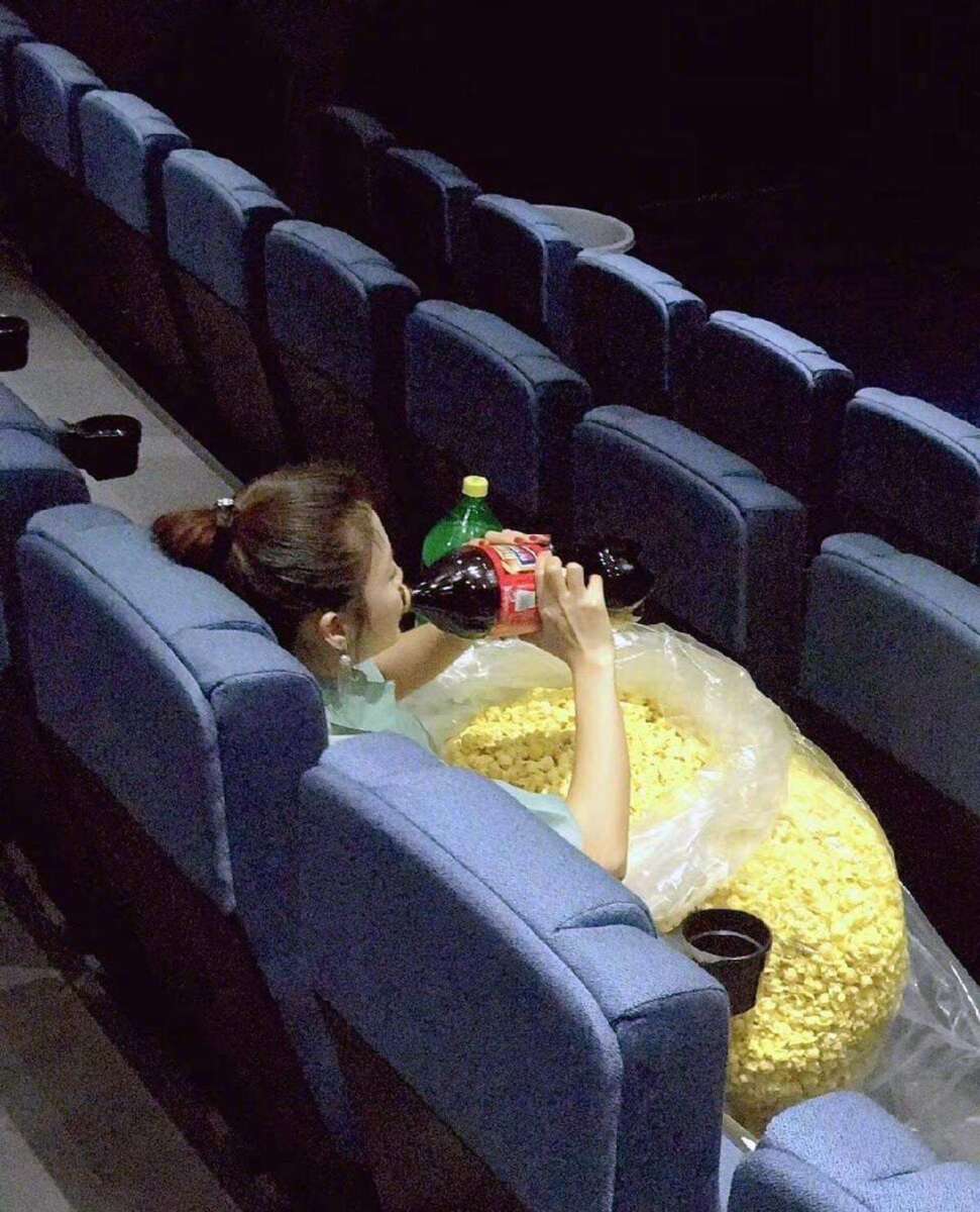 A woman sits alone in a movie theater, holding a giant plastic bag filled with popcorn and pouring soda from a large bottle into a cup, occupying several blue seats.