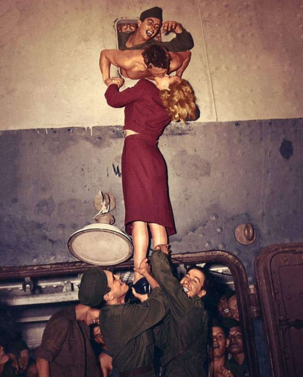 A group of soldiers lift a woman in a red dress so she can kiss a shirtless man leaning out of a hatch, while others watch and smile inside a crowded ship or train compartment.
