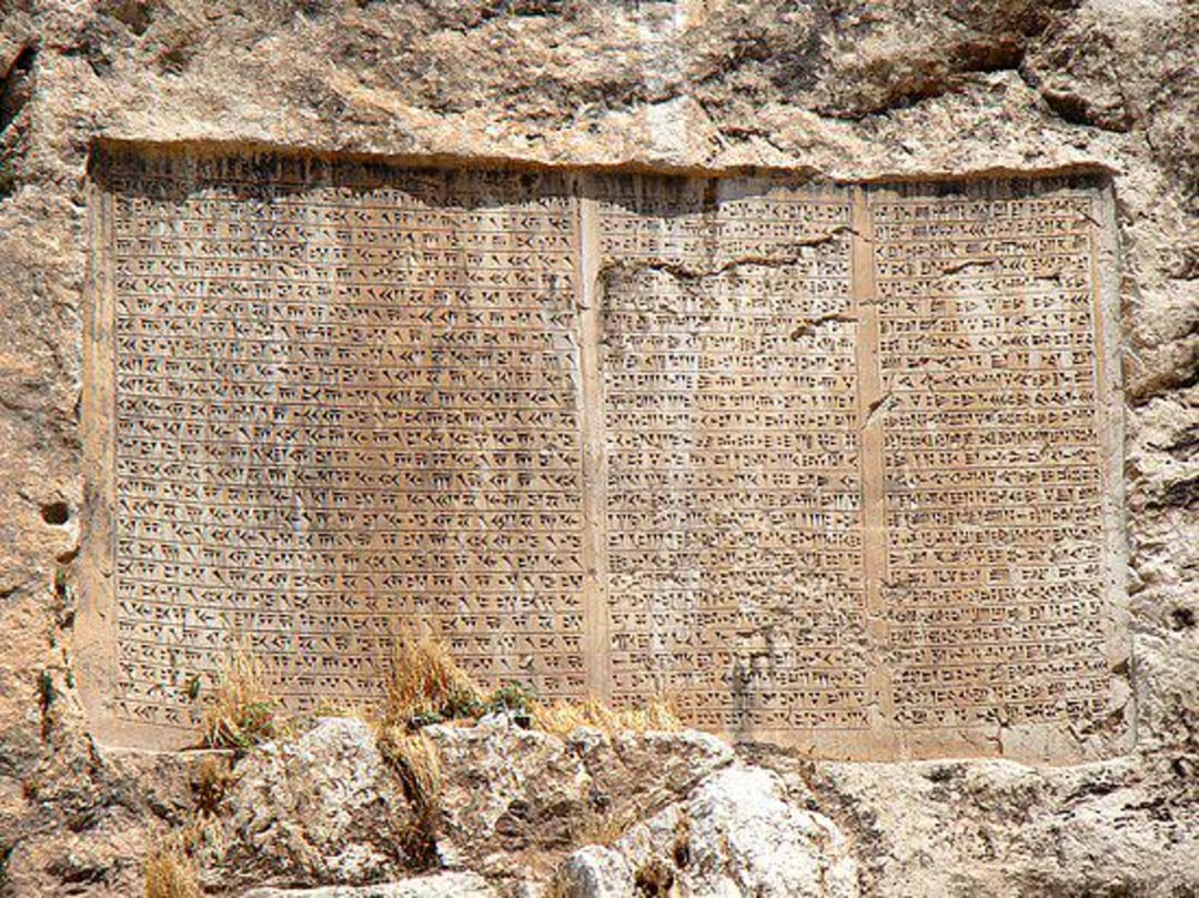 Ancient stone inscription carved into a rock face, featuring several lines of text in an old script, divided into panels. Tufts of dry grass grow at the base of the rocky surface.