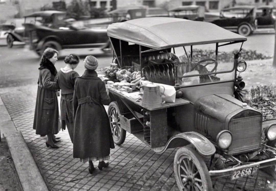 Three women stand at a vintage food truck parked on a city street, examining baked goods and other items displayed on the vehicle’s side. Old-fashioned cars are visible in the background.