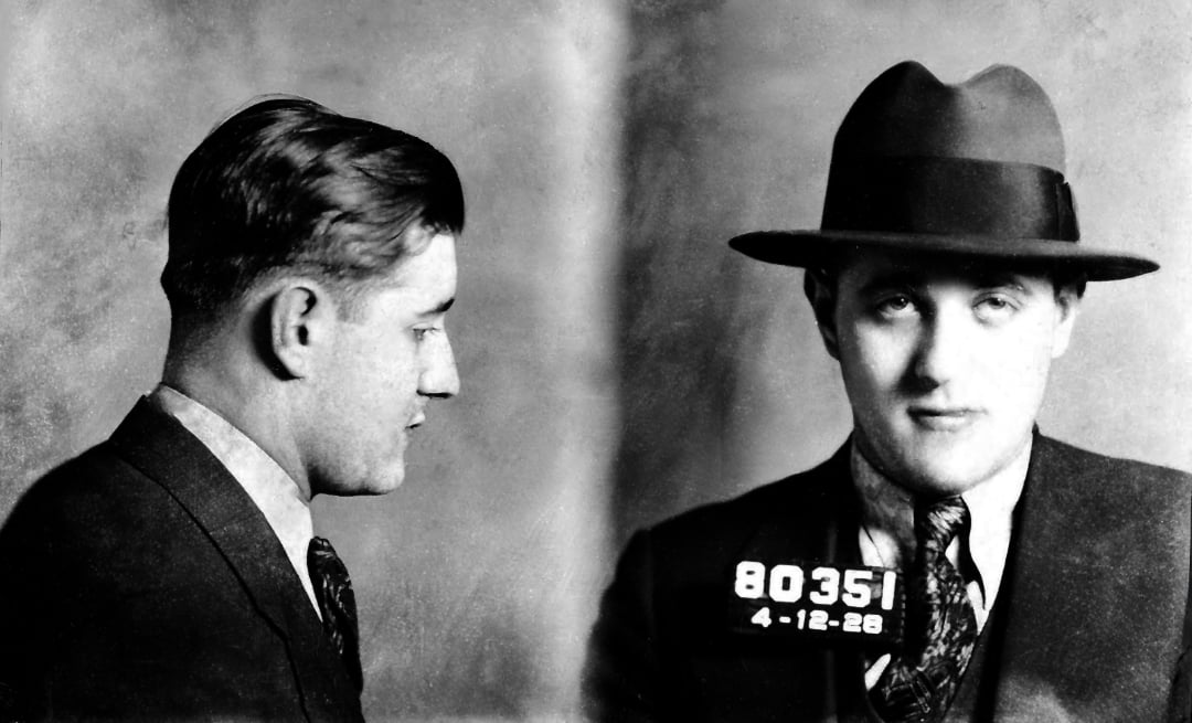 Black-and-white mugshot of a young man wearing a suit, tie, and fedora hat, shown in both profile and front view, holding a placard with the number 80351 and the date 4-12-28.