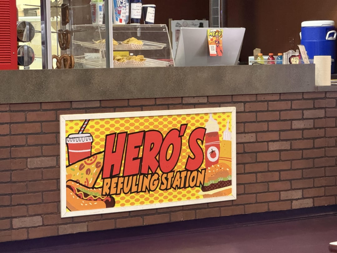 A counter with a sign reading "Hero's Refueling Station" in bold comic-style letters, featuring illustrations of a drink, hot dog, burger, and fries. Snacks and drinks are displayed on the countertop.