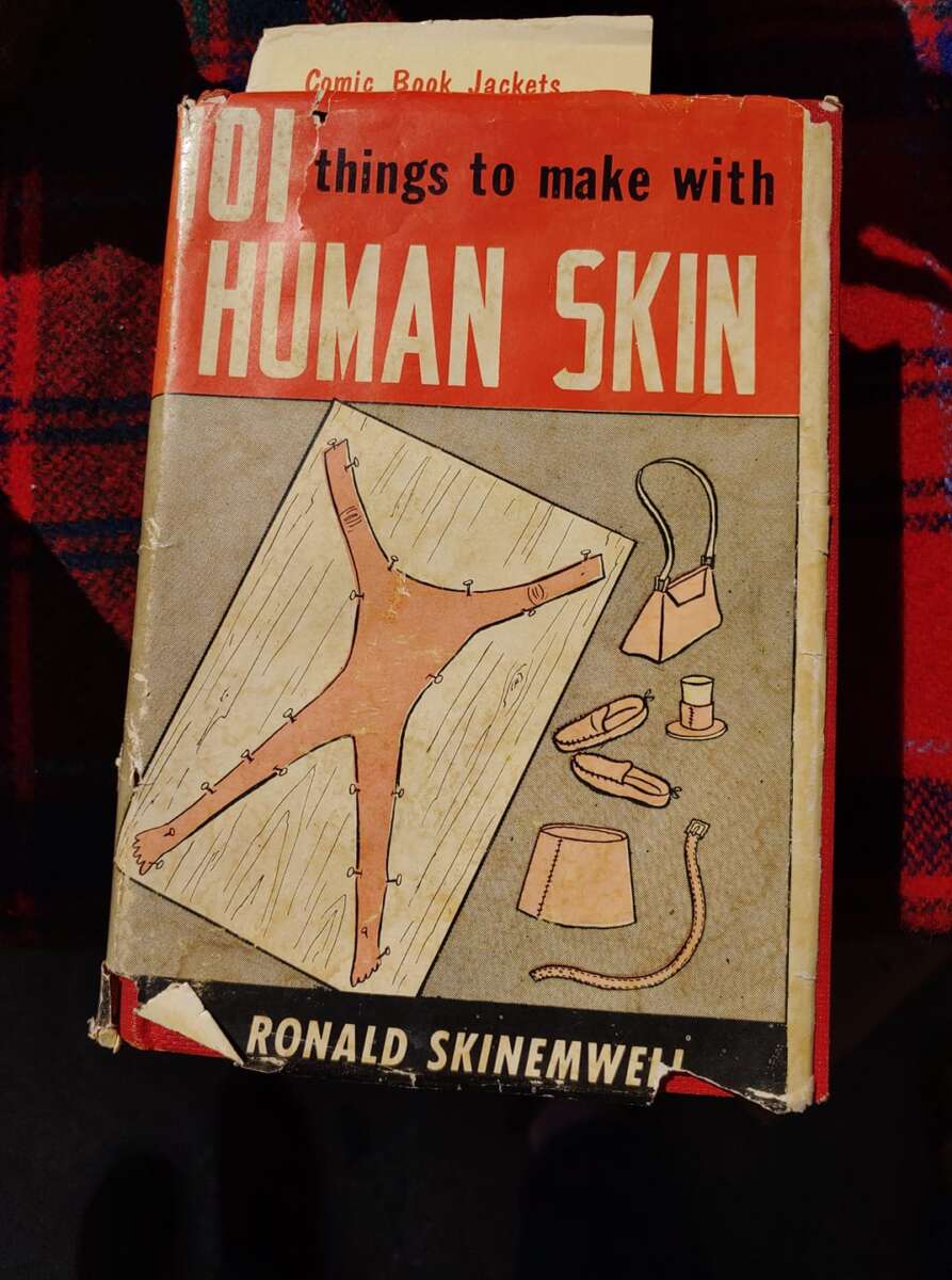 A worn book titled "101 Things to Make with Human Skin" by Ronald Skinemwell, with an illustration of stretched skin, tools, and small crafted items on the cover. A paper labeled "Comic Book Jackets" sticks out from the top.