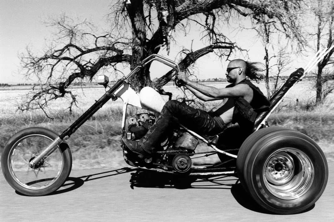 A person wearing sunglasses and a vest rides a custom chopper motorcycle with extended handlebars on a rural road, with bare trees and open fields in the background.