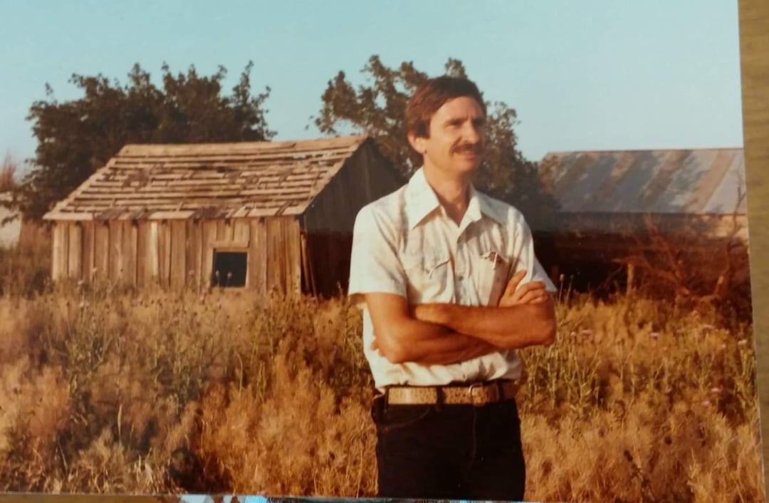 A man with a mustache, wearing a short-sleeve button-up shirt and jeans, stands with arms crossed in tall grass in front of two old wooden barns on a sunny day.