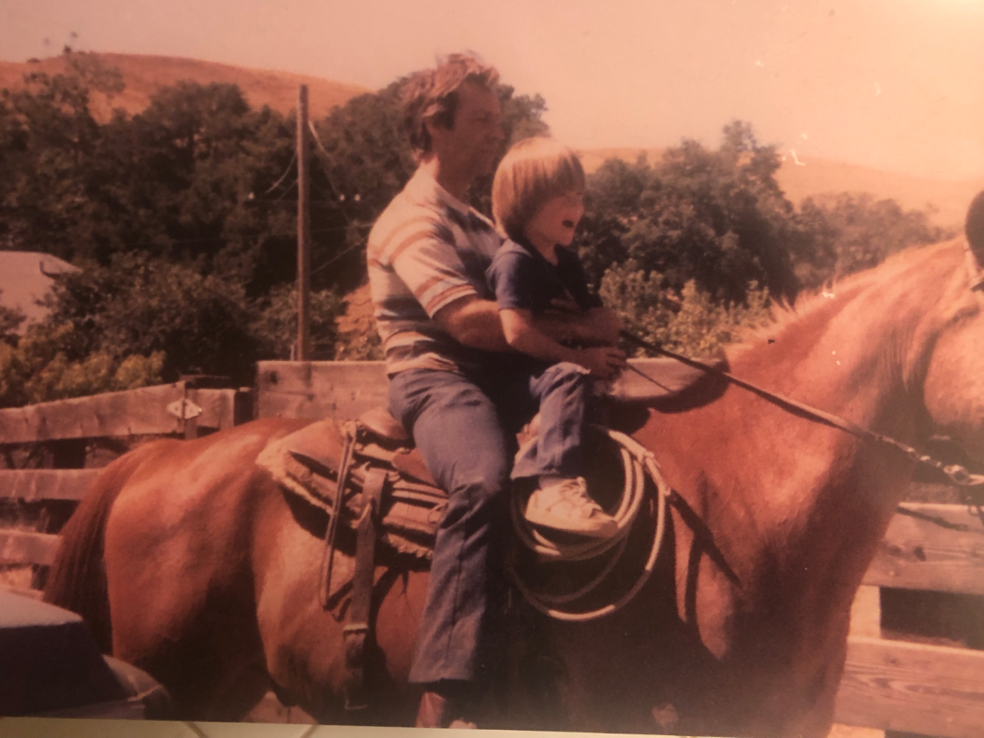 An adult and a young child sit together on a brown horse in a fenced outdoor area, with trees and hills in the background. The adult holds the reins while the child sits in front, both looking forward.