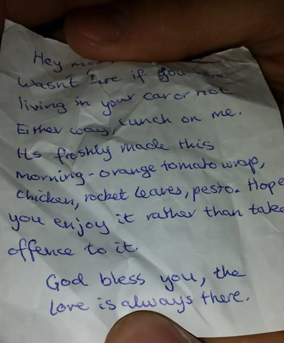 A handwritten note on crumpled white paper held between two hands. The message offers a meal and kind words, ending with "God bless you, the love is always there.
