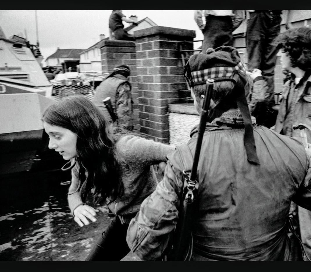 A black-and-white photo showing a young woman being apprehended by a soldier in military gear on a street, with other soldiers and an armored vehicle visible in the background. The scene appears tense and chaotic.