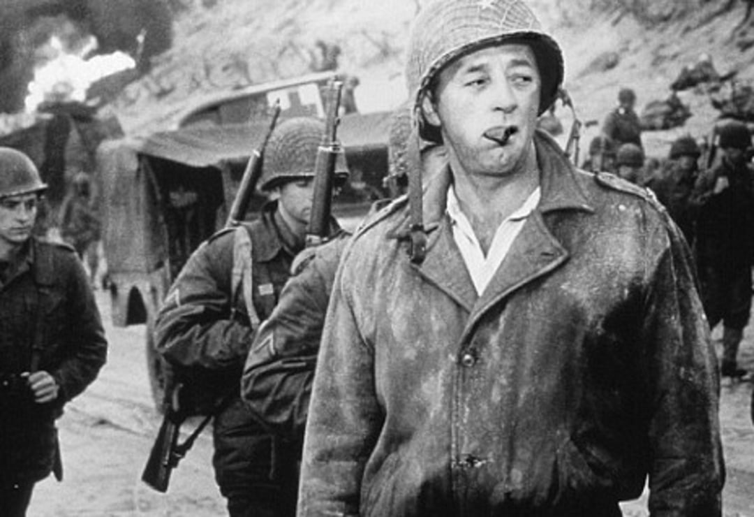 A soldier wearing a helmet and jacket smokes a cigar while walking on a beach. Other armed soldiers walk behind him, and a fire burns near vehicles in the background. The image is in black and white.