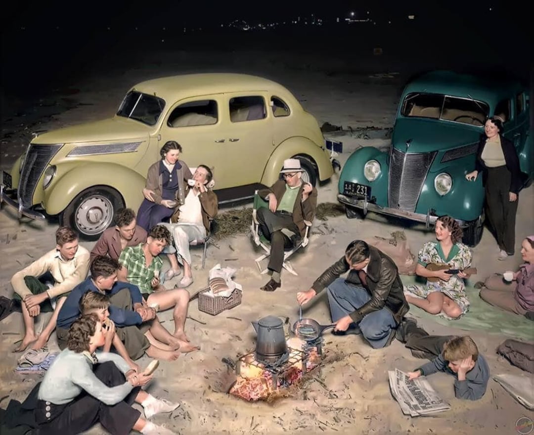 A group of people sit around a campfire on a beach at night, with two vintage cars parked behind them. Some are talking, others are reading, and there are baskets and a pot over the fire.