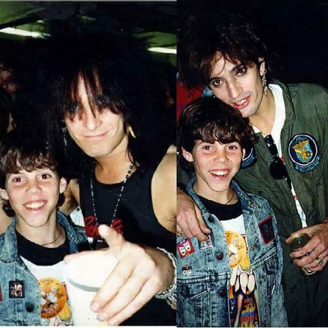 Two young men in denim jackets pose with rock musicians at an event. One musician has dark, wild hair and a tank top, while the other wears a green flight suit and holds a drink. All are smiling for the camera.