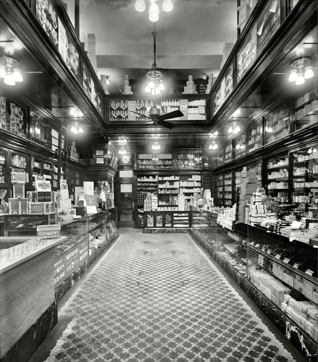 Black and white photo of an old-fashioned grocery store with shelves lining the walls, filled with assorted boxes and goods. Glass display cases hold various products. Ornate lights hang from the ceiling above a patterned tile floor.