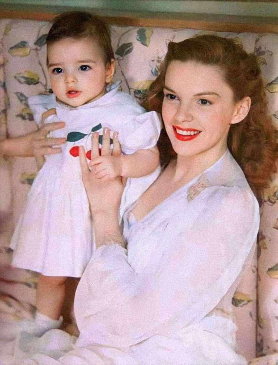 A smiling woman with wavy brown hair and red lipstick holds a baby wearing a white dress with a cherry design. They are sitting on a patterned couch with a soft, pastel floral print.