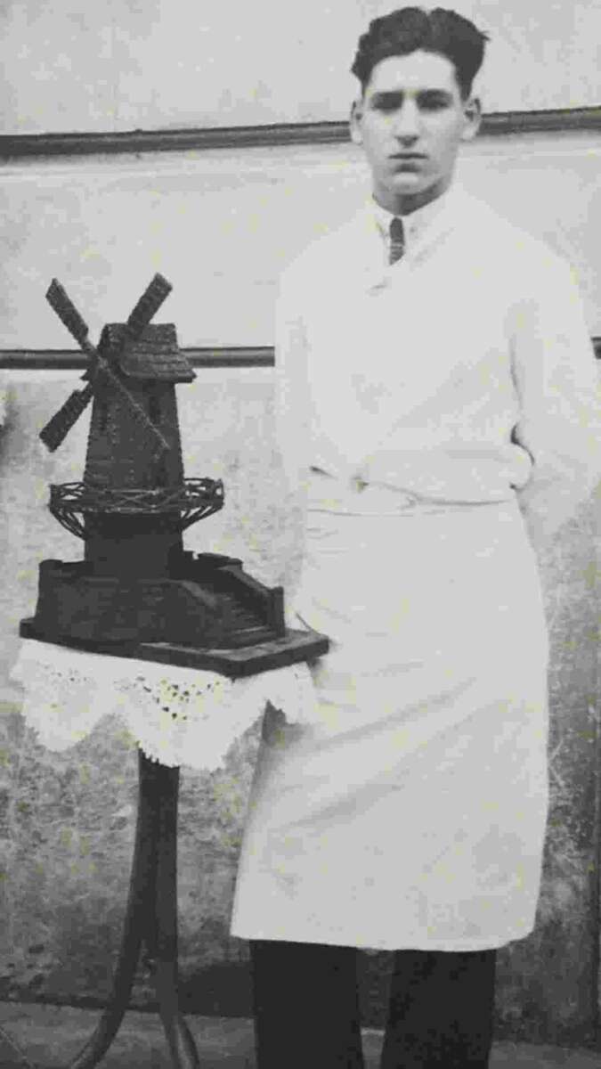 A young man in a white apron stands beside a small table displaying a detailed model of a windmill, possibly made of chocolate, on a lace-covered cloth. The background is a plain, textured wall.