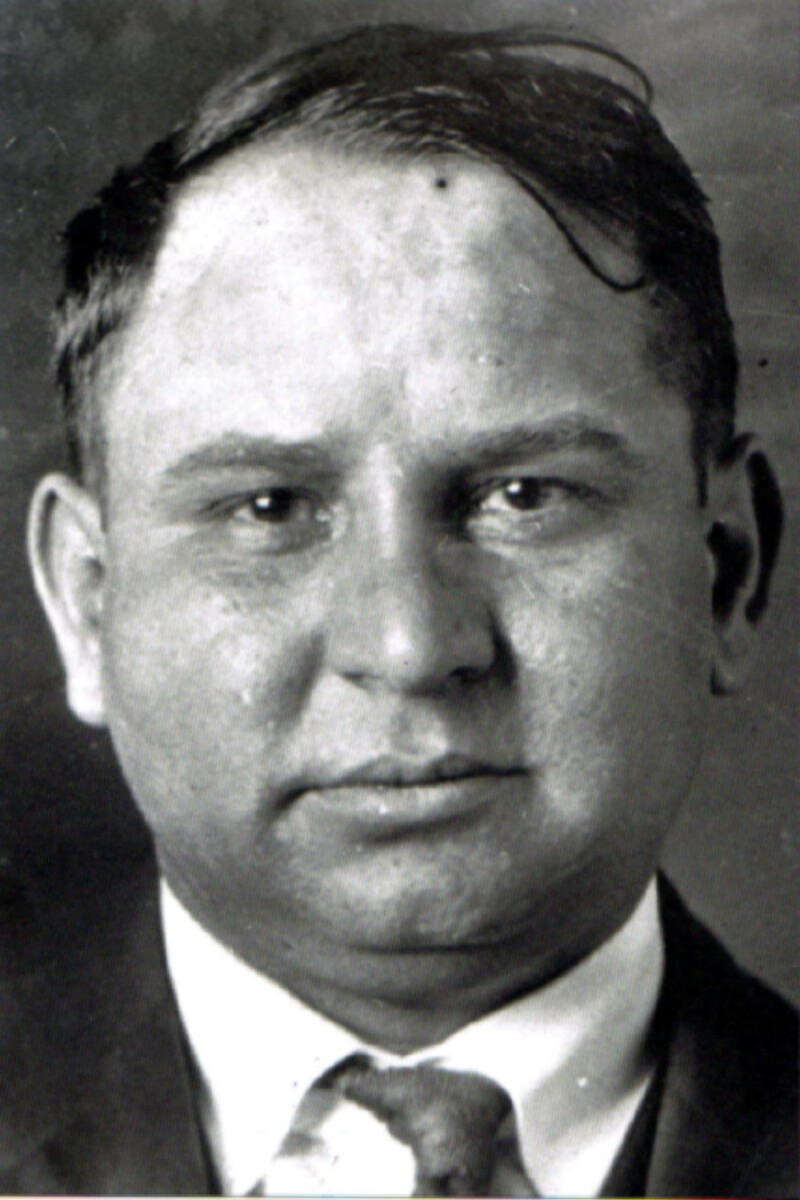 Black and white portrait of a man with short dark hair, wearing a suit and tie, looking directly at the camera with a neutral expression.